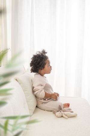 A small child in beige clothing lies on a white couch near a window with soft natural light and green plants.