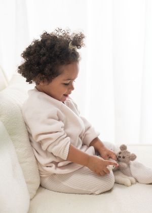 Baby in cream-colored sweater sits on white bedding while playing with a small stuffed toy.