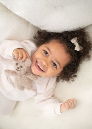 A child with curly hair lies on a white surface while holding a small stuffed animal toy.