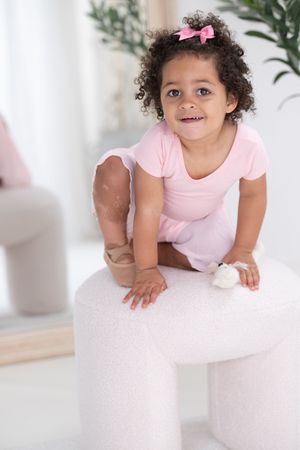 A young child in a pink shirt and bow plays on a white carpet in a bright room with plants in the background.