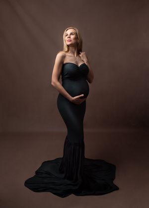 A graceful figure in a flowing black evening gown poses elegantly against a dark brown studio backdrop.