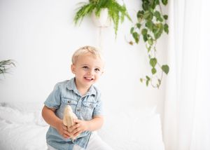 Baby in denim outfit sits on white floor with green plants hanging in background.