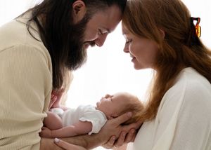 A bearded father and red-haired mother lovingly cradle their newborn baby together in a tender family portrait.