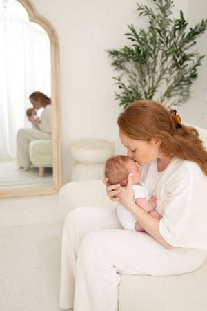A red-haired mother gently kisses her newborn baby while seated on a white chair, with a mirror reflection in the background.