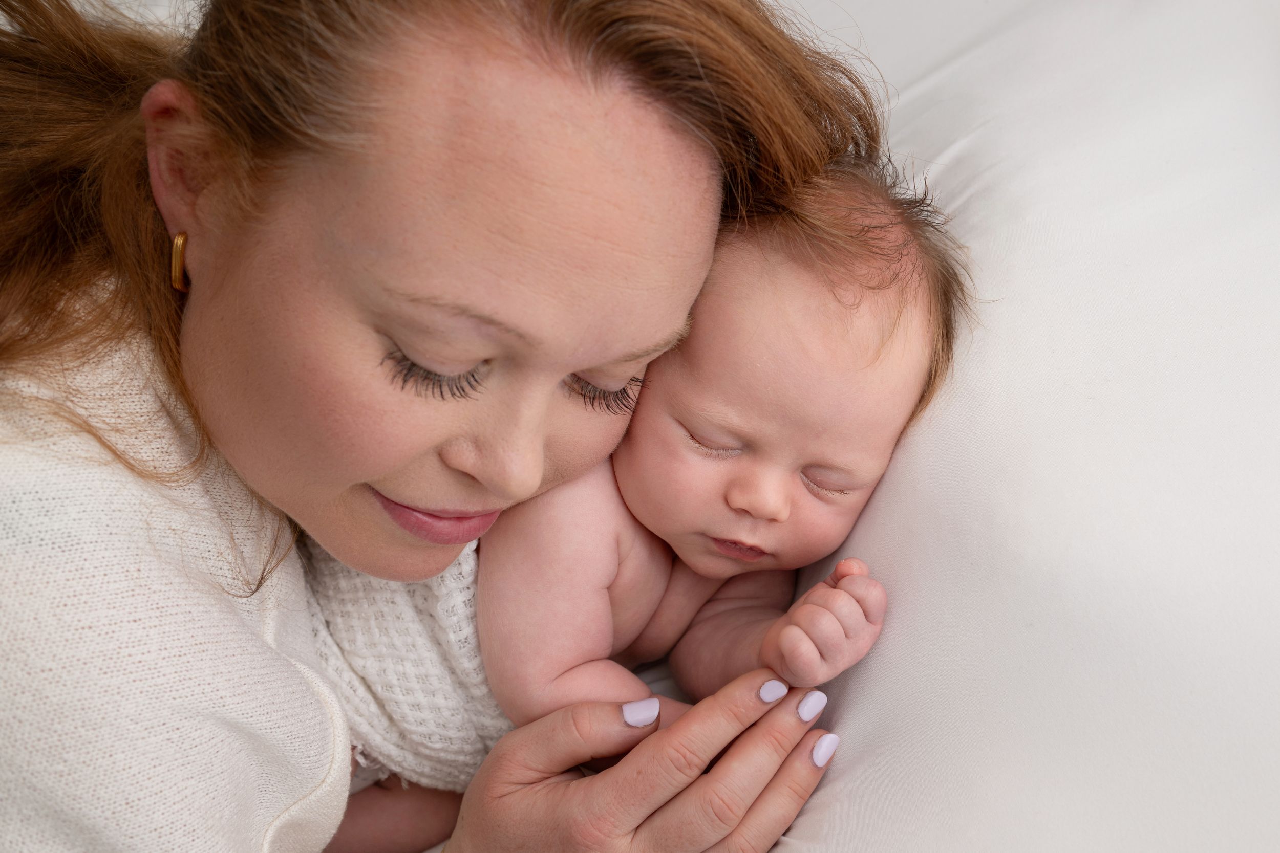 A mother with red hair tenderly holds her sleeping newborn baby close to her face on a white background.