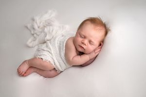 Peaceful newborn baby sleeping on white background wearing a white knit romper with soft natural lighting.