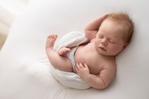 Peaceful newborn baby with red hair sleeping curled up on white blanket, wrapped in knit fabric.