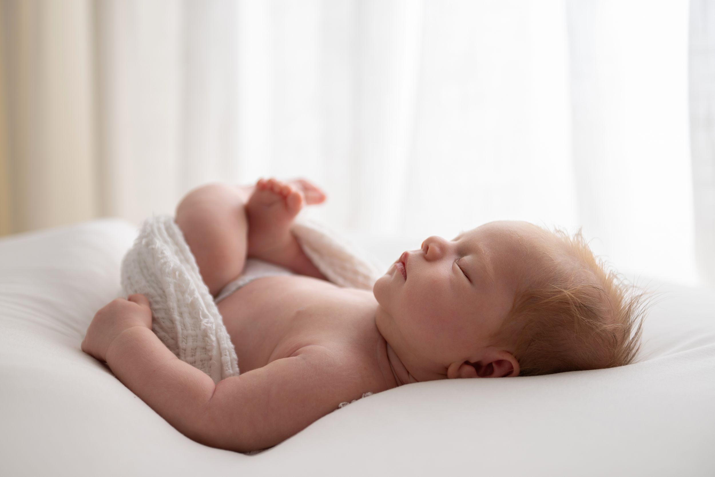 Peaceful newborn baby sleeping on white bedding in soft natural light, wearing a diaper.