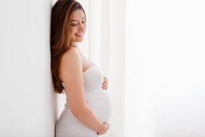 Elegant maternity portrait with woman in white dress against bright window backdrop.