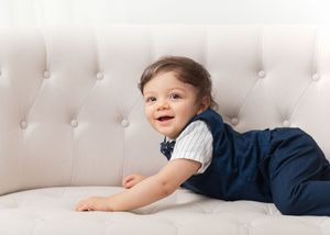 Baby in white shirt and navy pants lying on tufted white couch while playing.