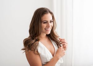 A woman with wavy brown hair smiles while wearing a white lace top near a bright window.