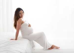 Maternity photoshoot in bright white room with soft natural lighting showing pregnant woman in white dress.