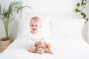 A baby sits on white bedding with a teddy bear while palm plants decorate the bright, airy room.