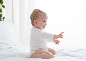 Happy baby in white onesie sits on white bedding while reaching out and laughing.