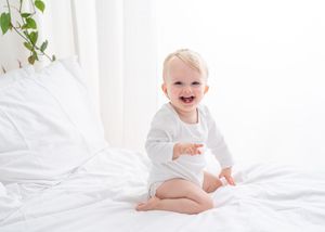 A baby in a white onesie sits and laughs joyfully on white bedding while reaching towards the camera.