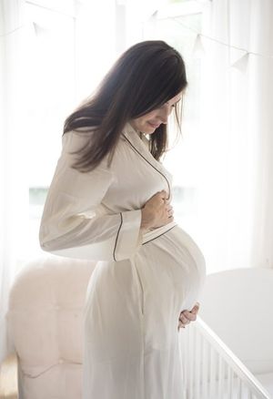 Pregnant woman in white dress standing by window in soft natural light creating ethereal maternity portraits.