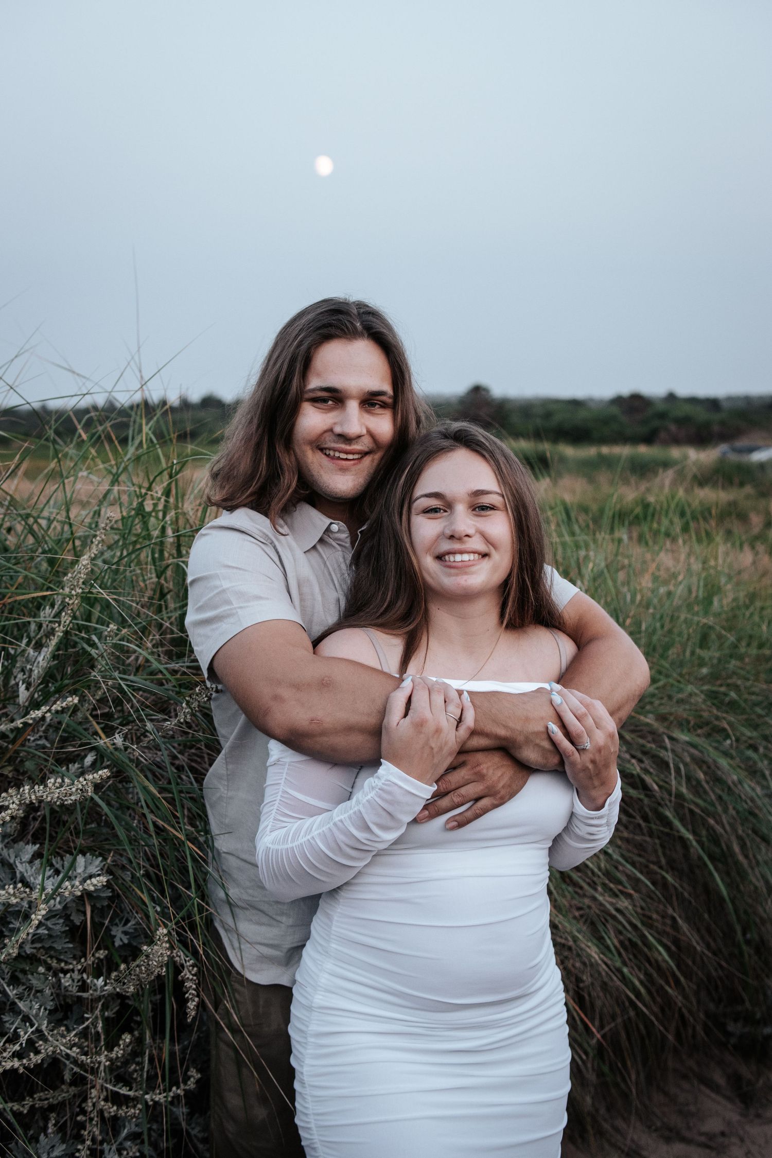 A couple embraces in a grassy field at dusk with the moon visible in the background.