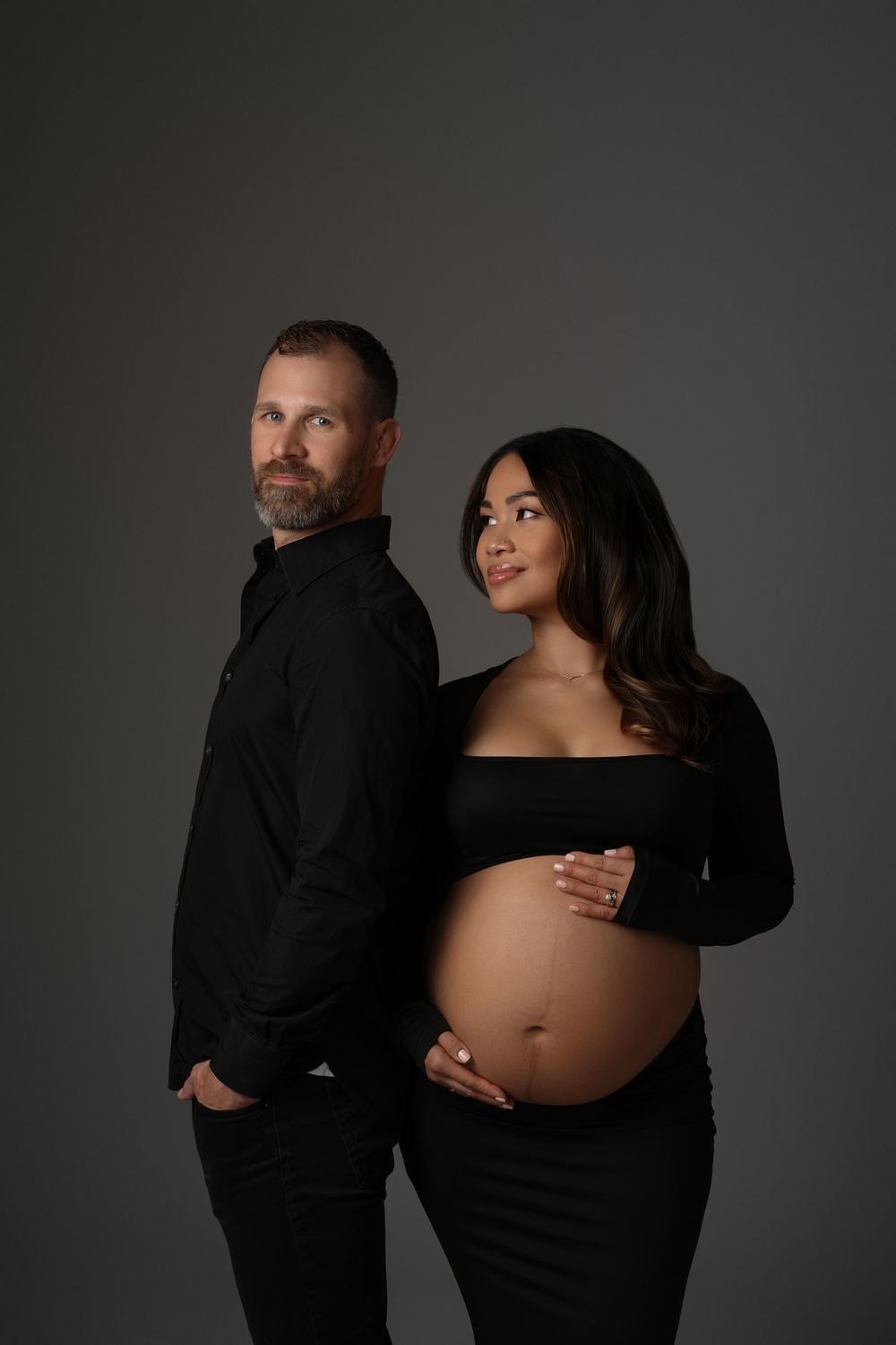 Pregnant woman and bearded man pose back-to-back in black outfits in studio.