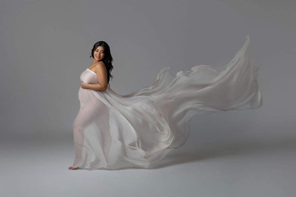 Pregnant woman in white draped fabric gown with flowing train poses in a dreamy maternity studio photo.