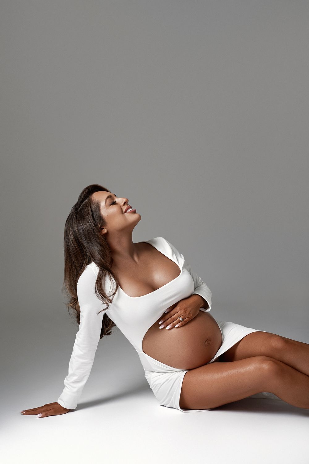 Pregnant woman in white crop top sitting on floor with eyes closed in a gray studio.