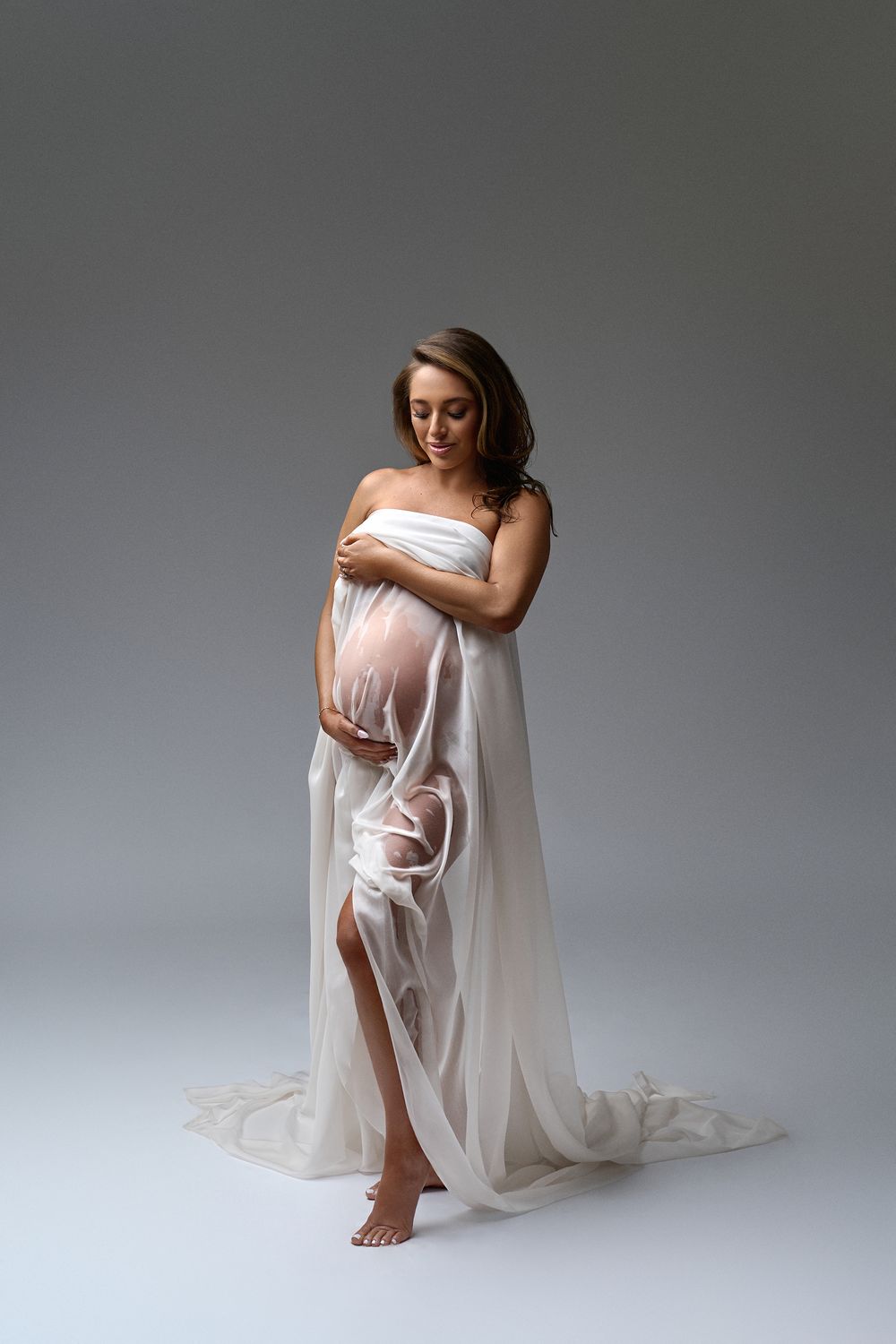 Pregnant woman in flowing white chiffon wrap gown cradling her baby bump in a studio.
