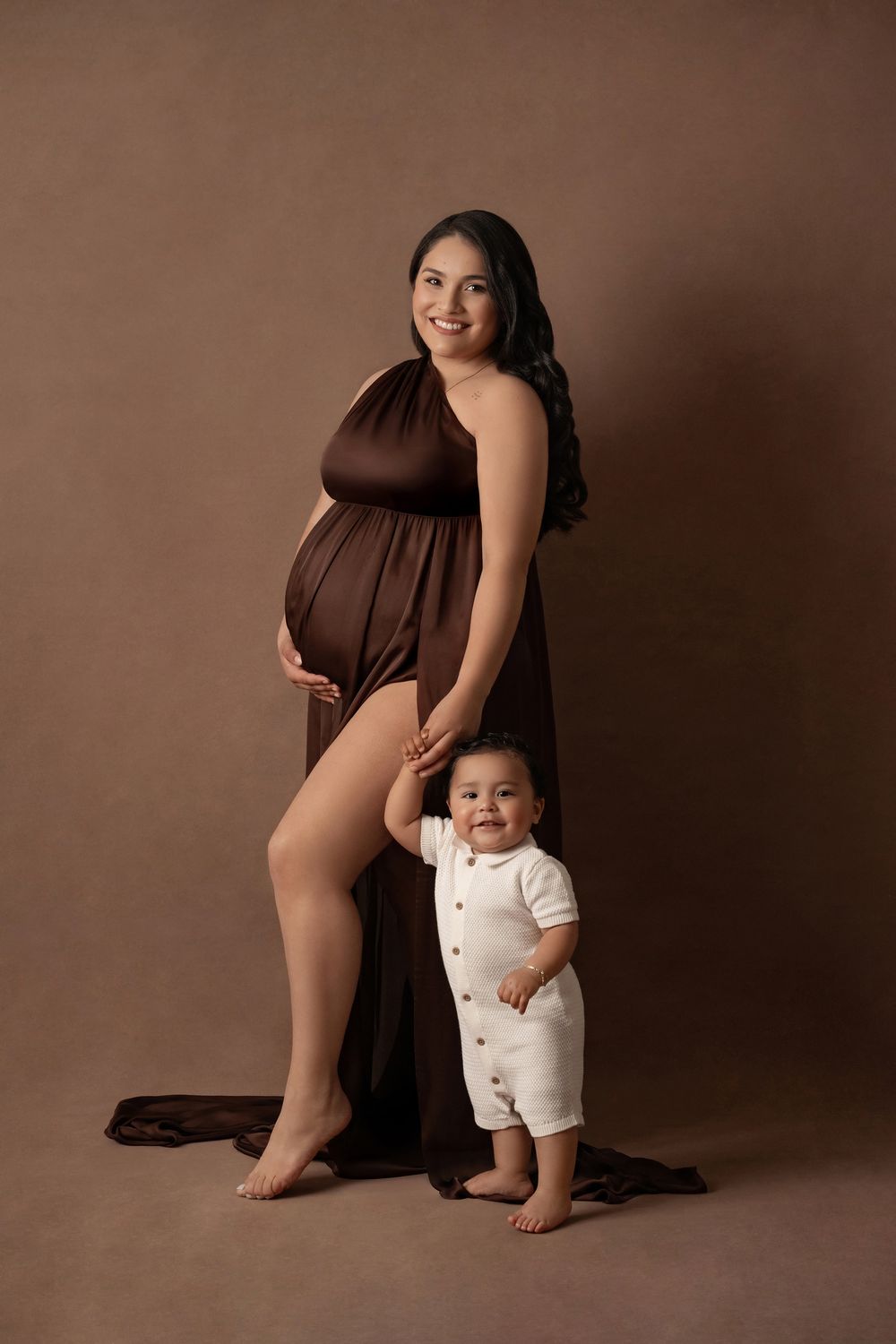 Pregnant woman in brown draped gown poses with smiling toddler in white romper during maternity studio session.