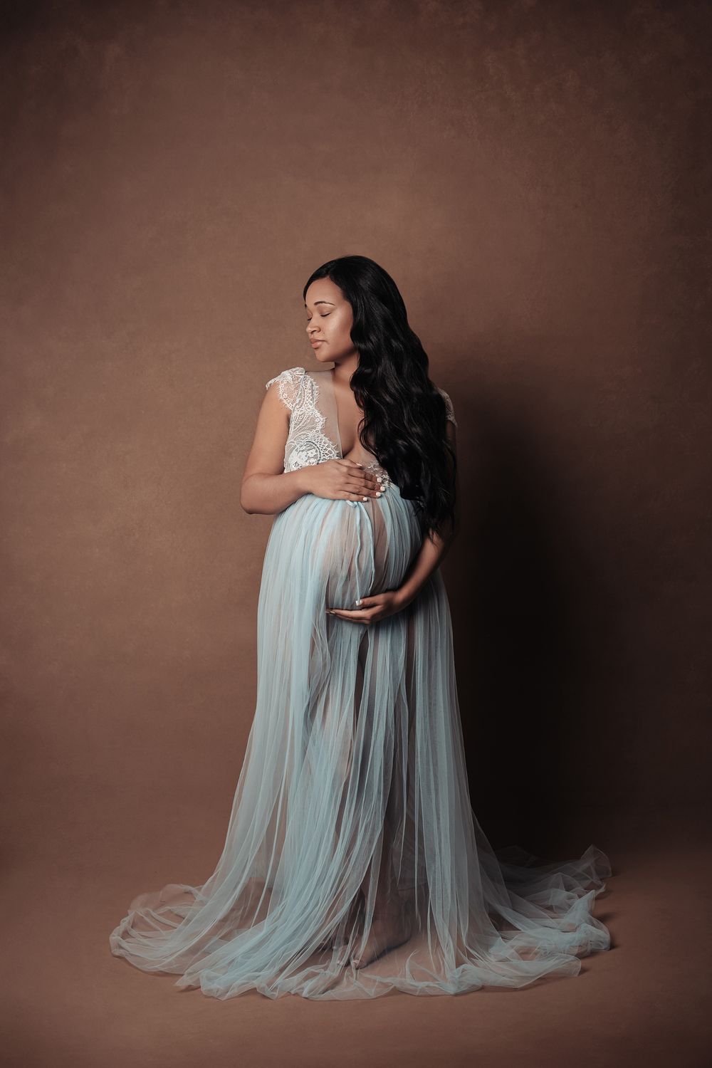 Pregnant woman in white lace and blue tulle gown poses in brown studio.