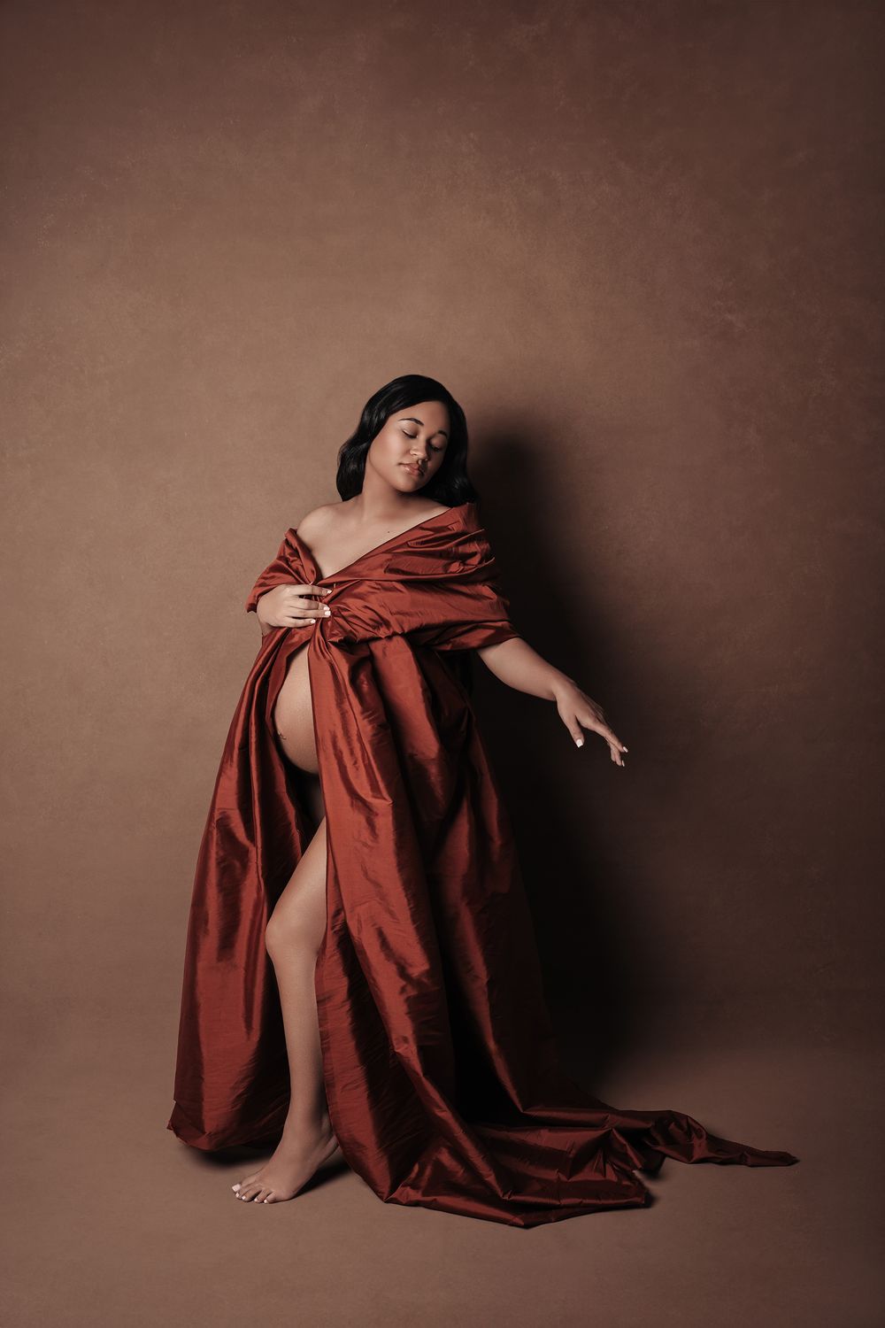 Pregnant woman draped in flowing rust satin fabric poses in brown studio.