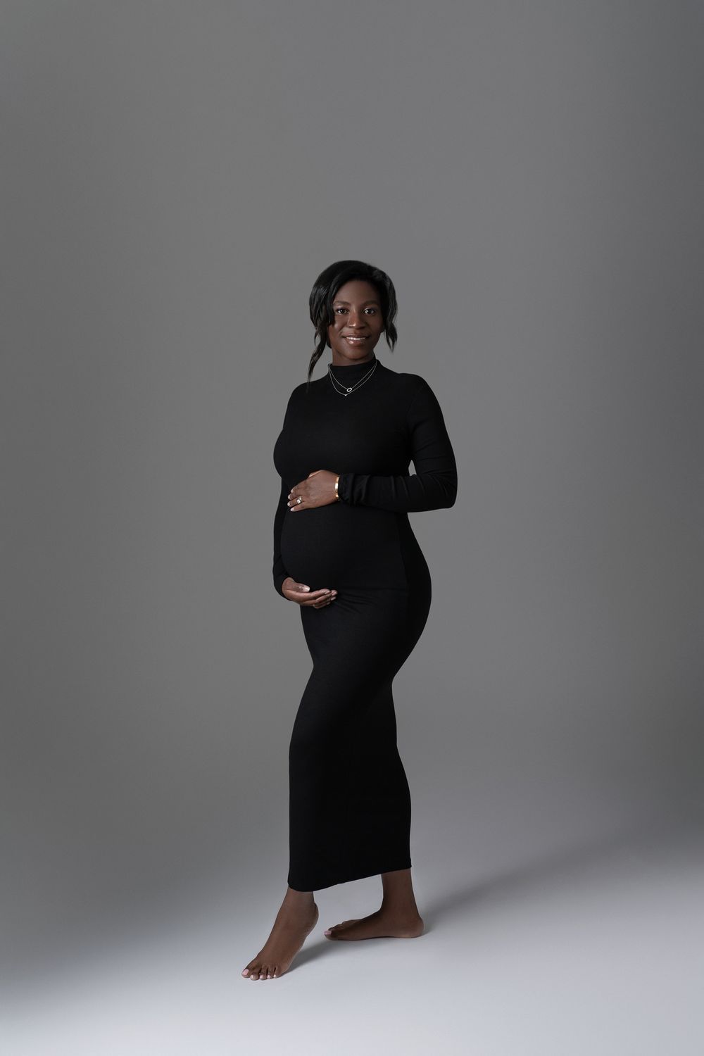 Smiling pregnant woman in fitted black long-sleeve dress cradles her baby bump in a gray studio setting.