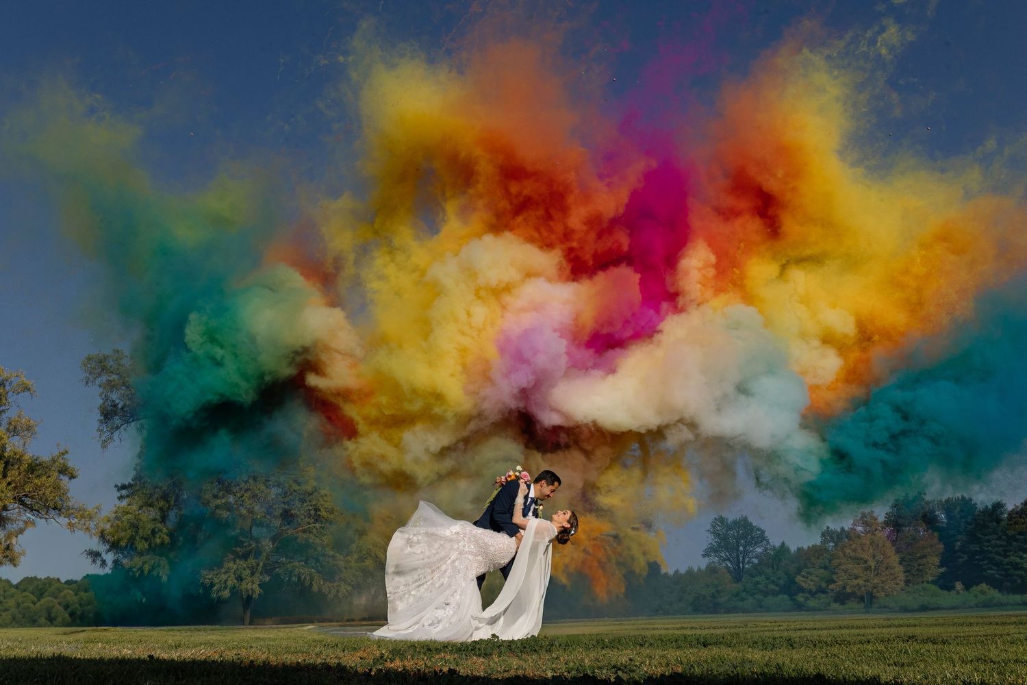 A couple embraces in a white dress against a dramatic backdrop of colorful smoke bombs in vibrant rainbow hues.