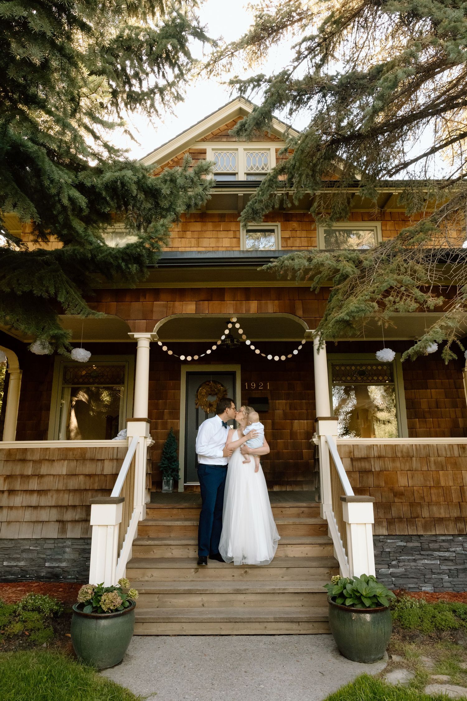 A rustic cedar-shingled craftsman house with white columns and string lights decorating the front porch entrance.