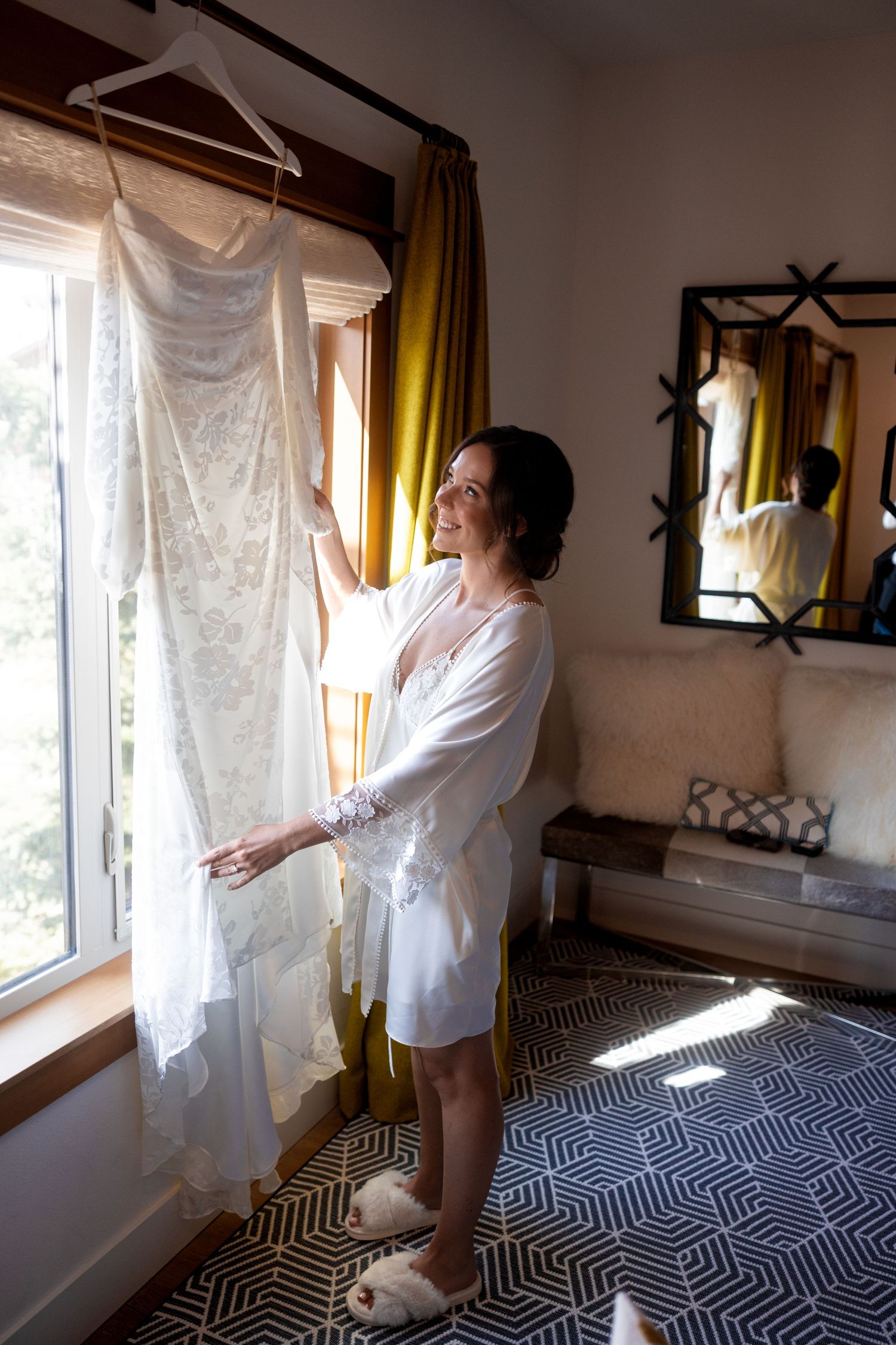 Person in white robe admiring wedding dress hanging by window with yellow curtains.
