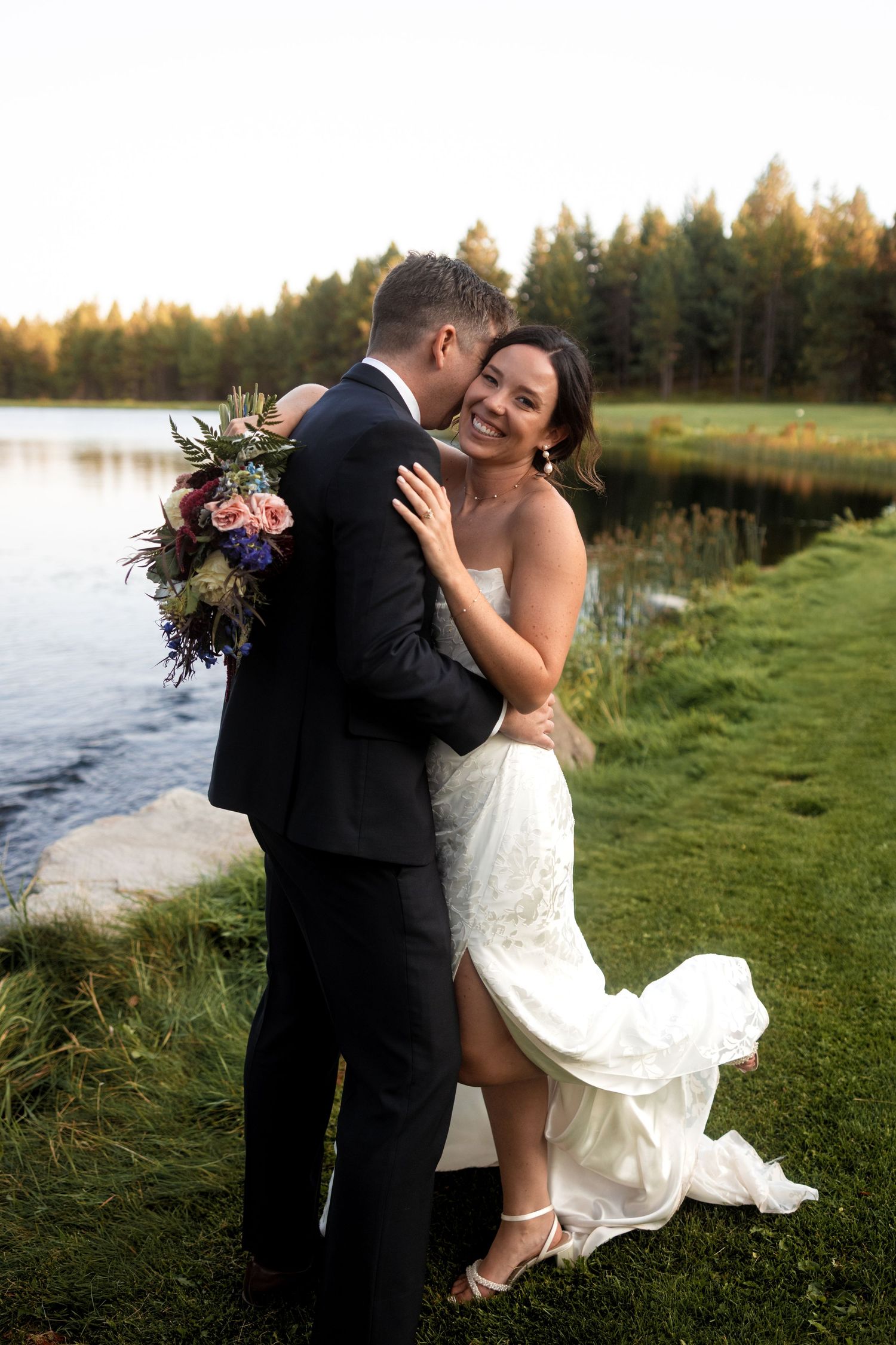 Romantic couple embraces on grassy waterfront during sunset wedding photoshoot.