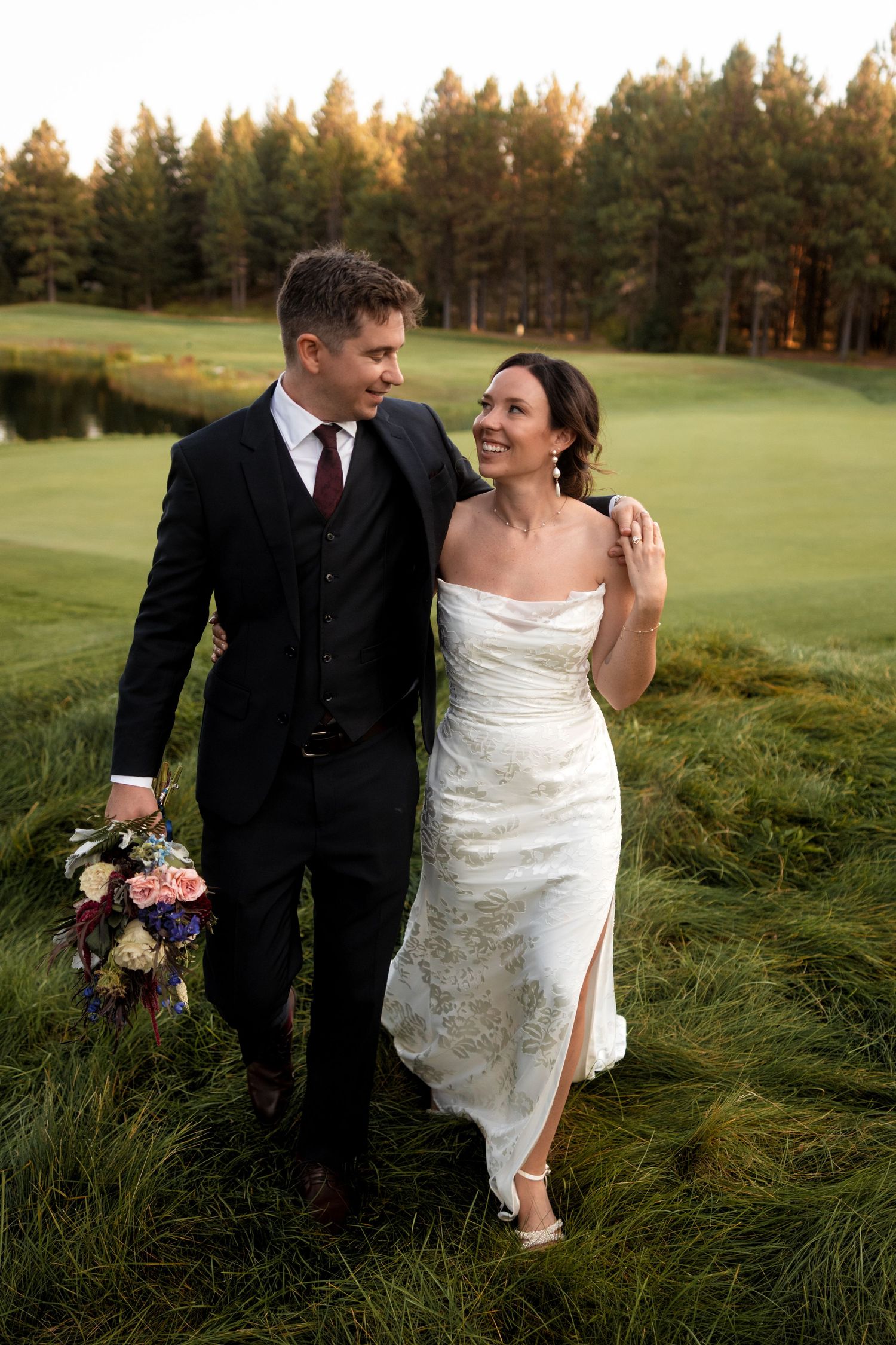 Wedding couple walking through tall grass with evergreen trees in background.