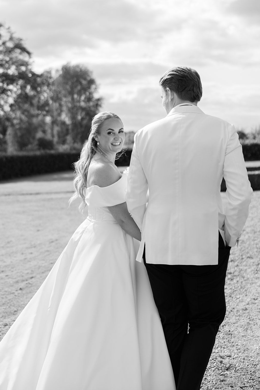 A romantic black and white wedding photo of a couple outdoors, with the bride in a flowing white gown looking back at camera.