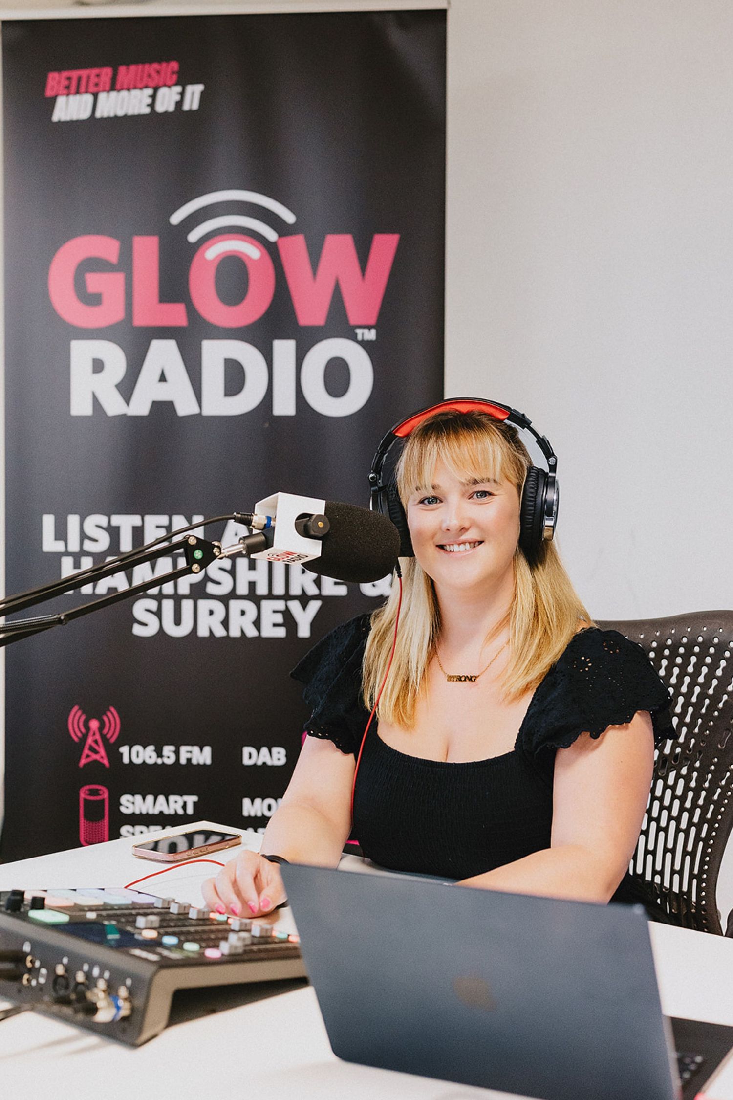 Radio host wearing headphones sits at broadcasting desk with laptop and equipment in professional studio setting.