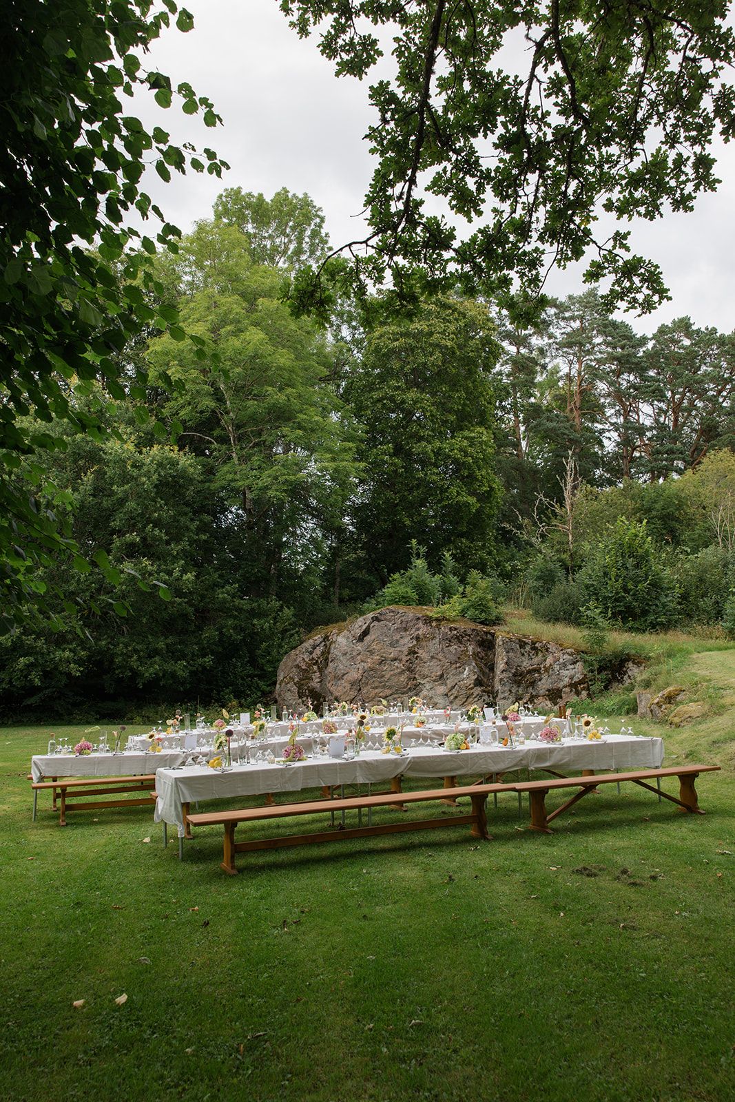 Long wooden banquet tables with place settings arranged outdoors on a lush green lawn beside a rocky outcrop.