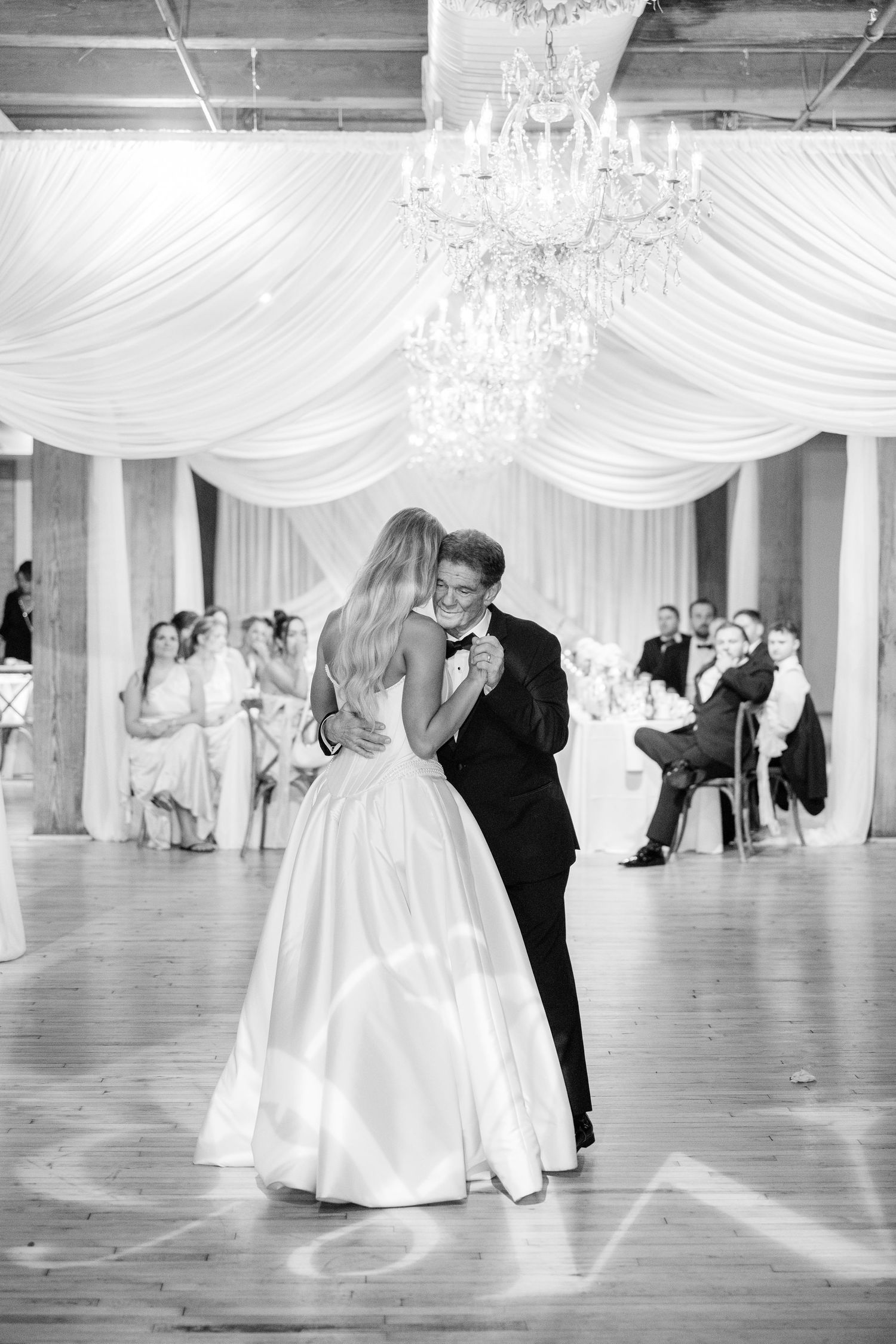 Couples dance gracefully across an elegant ballroom with white draped ceiling and crystal chandeliers.
