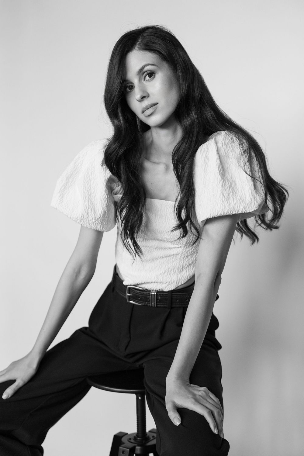 Black and white fashion portrait of someone in a white puff sleeve top and dark pants sitting on a stool.