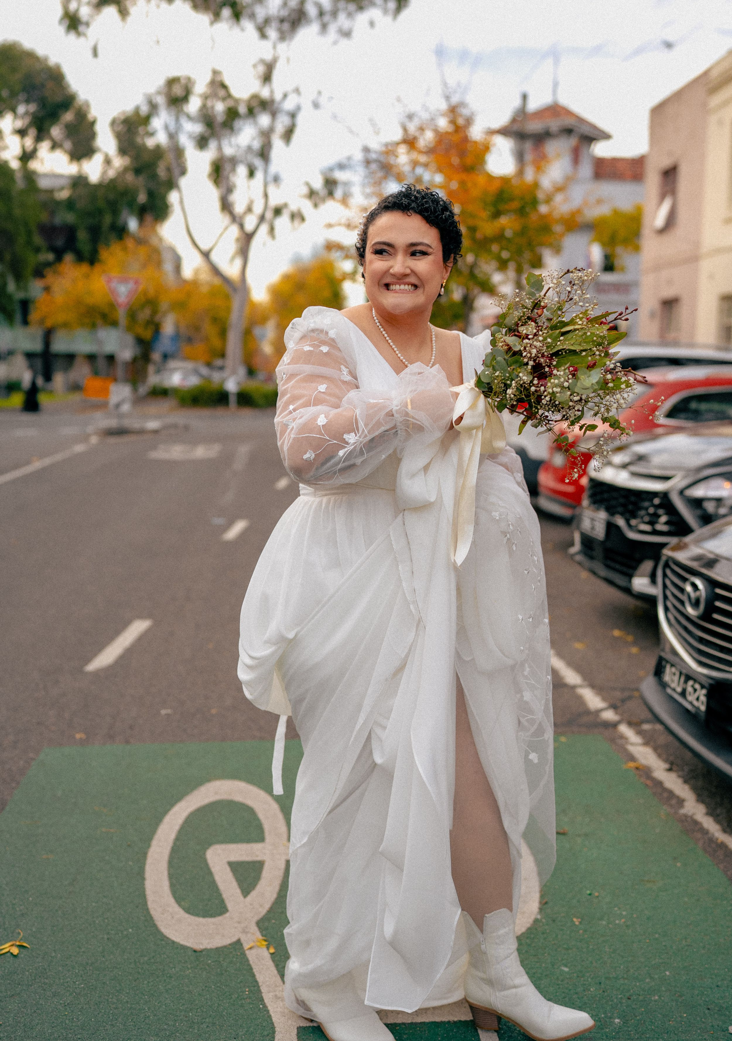 A person in a flowing white wedding dress and boots stands on a bike lane holding a bouquet on an autumn day.