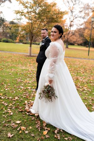 A couple in wedding attire dances in a park during autumn with fallen leaves scattered on the ground.