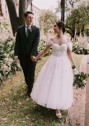 A bride in a tea-length tulle wedding dress and groom in dark suit hold hands while walking through a garden setting.