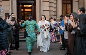 Wedding guests line up outside a historic stone building to watch a newlywed couple exit during their celebration.