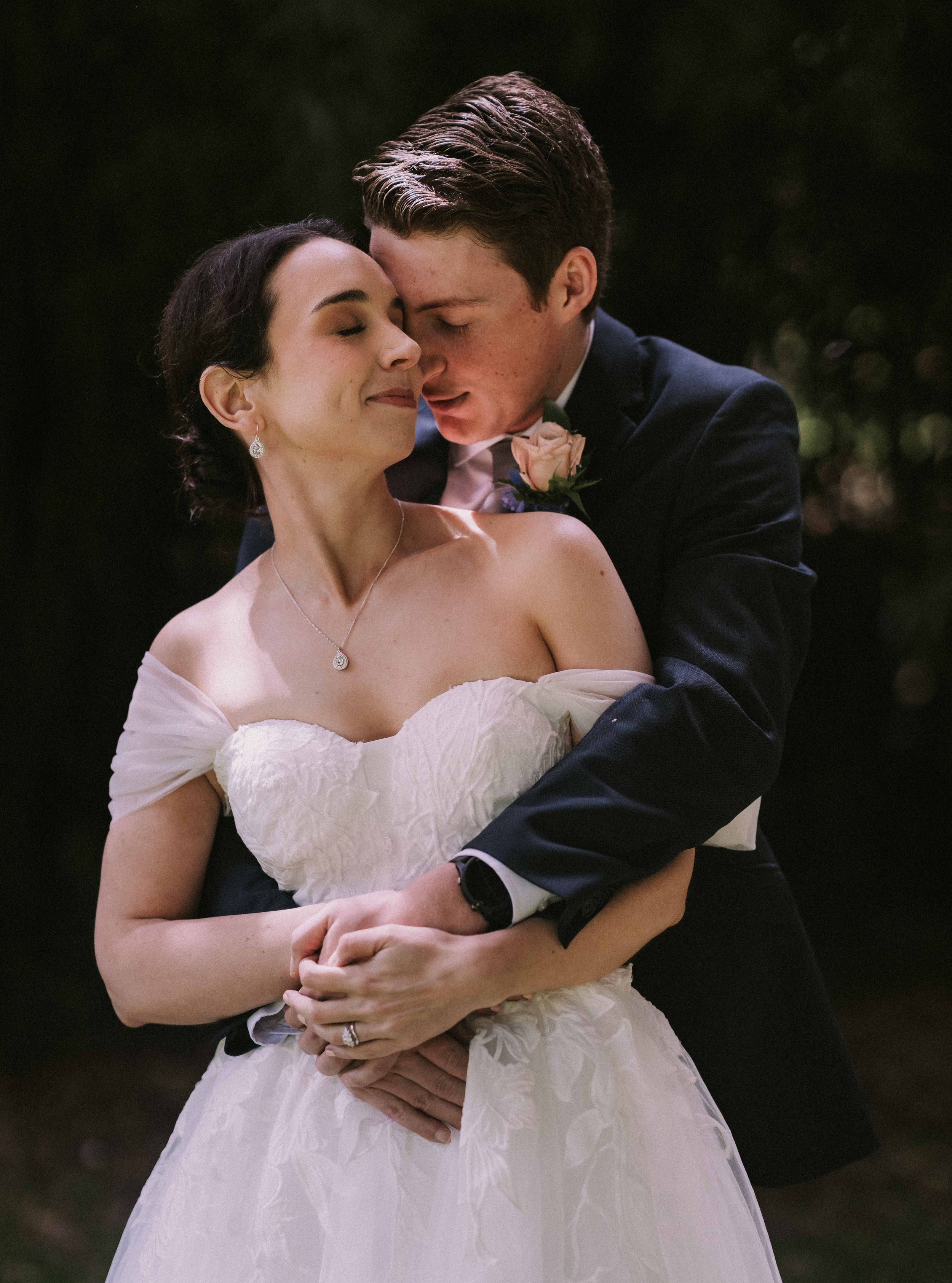 A romantic wedding portrait shows a couple embracing in soft natural lighting, with the bride in a white strapless gown.