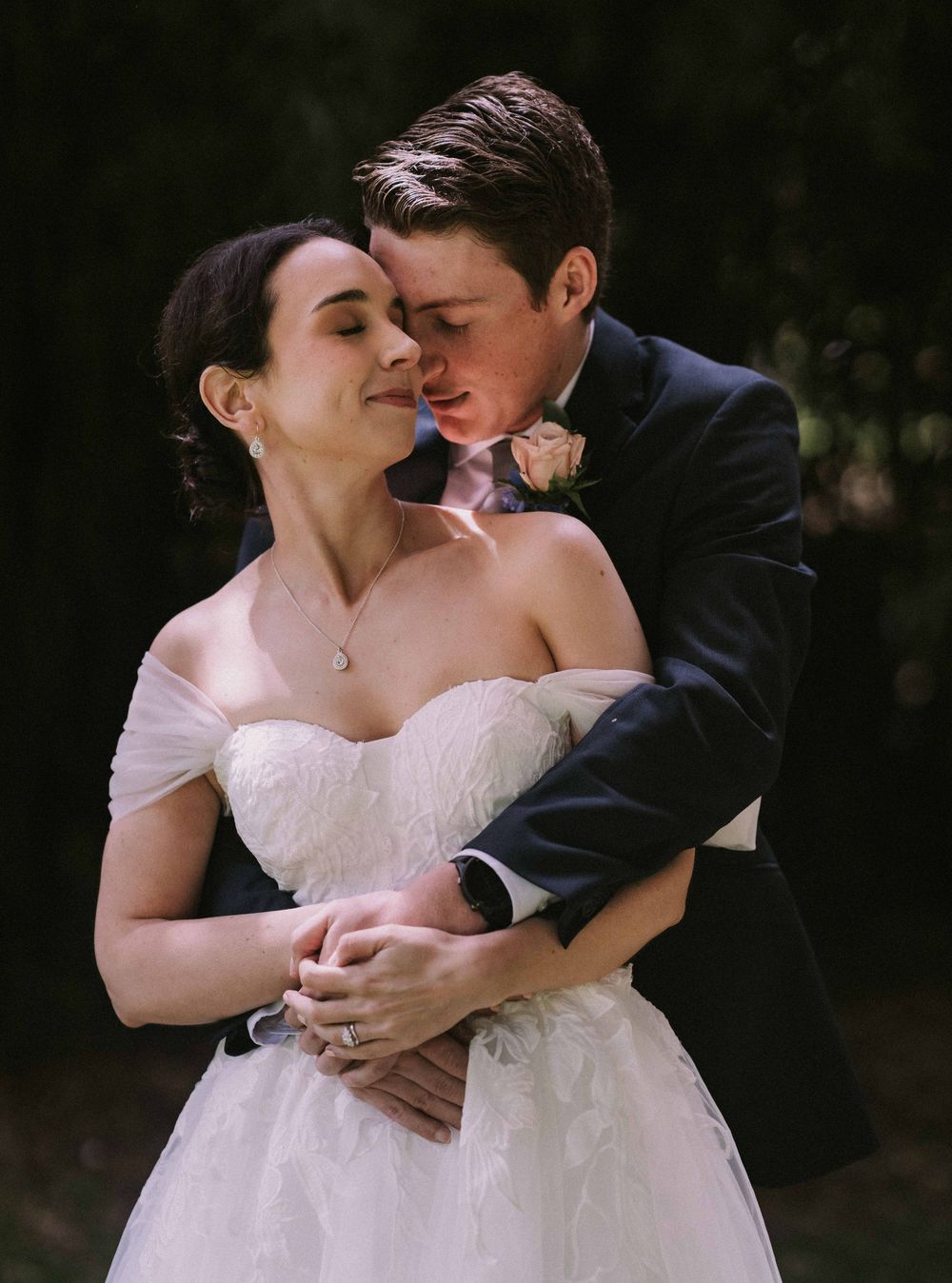 A romantic wedding portrait shows a couple embracing in soft natural lighting, with the bride in a white strapless gown.