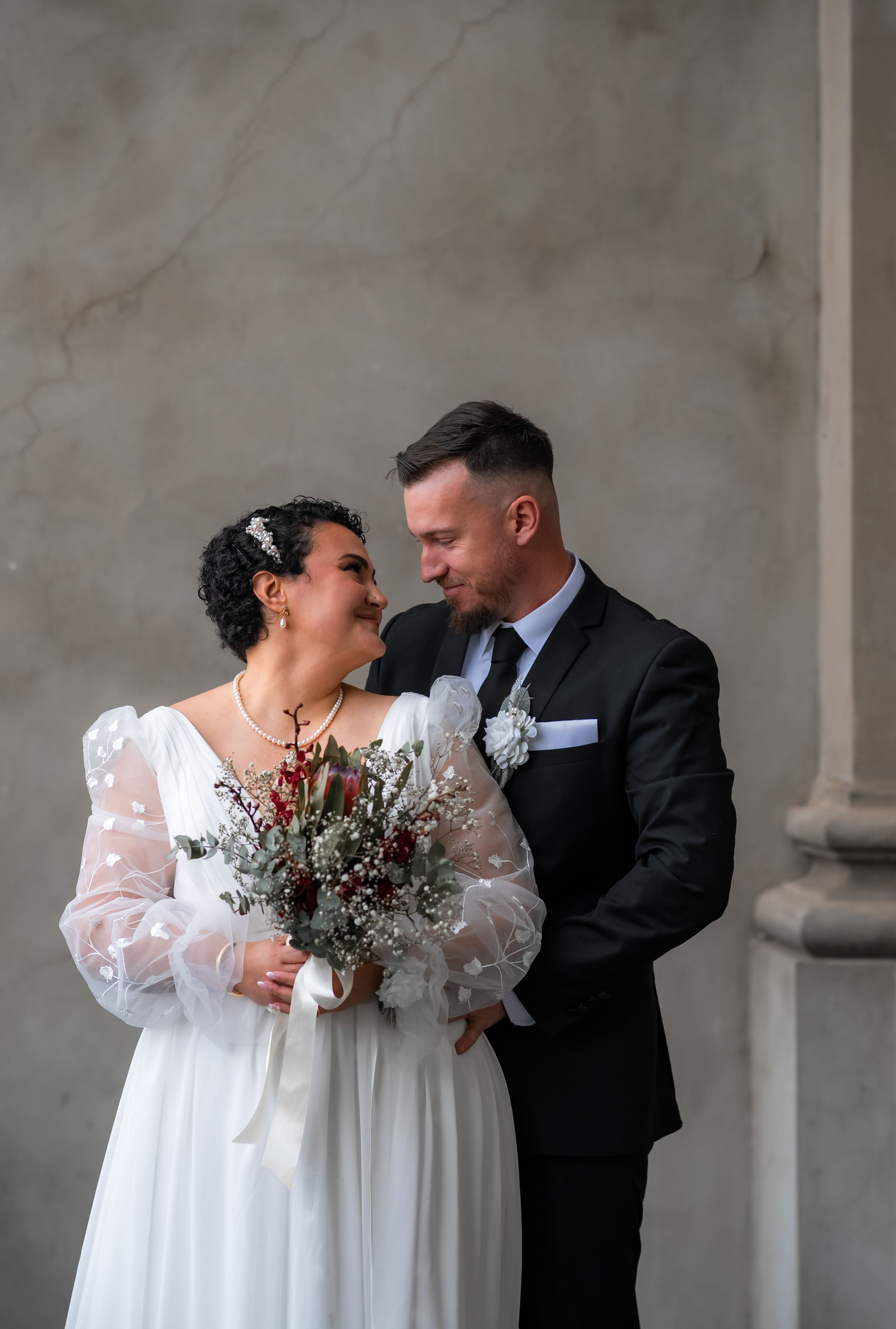 A couple in wedding attire share an intimate moment against a grey stone wall in a romantic outdoor setting.