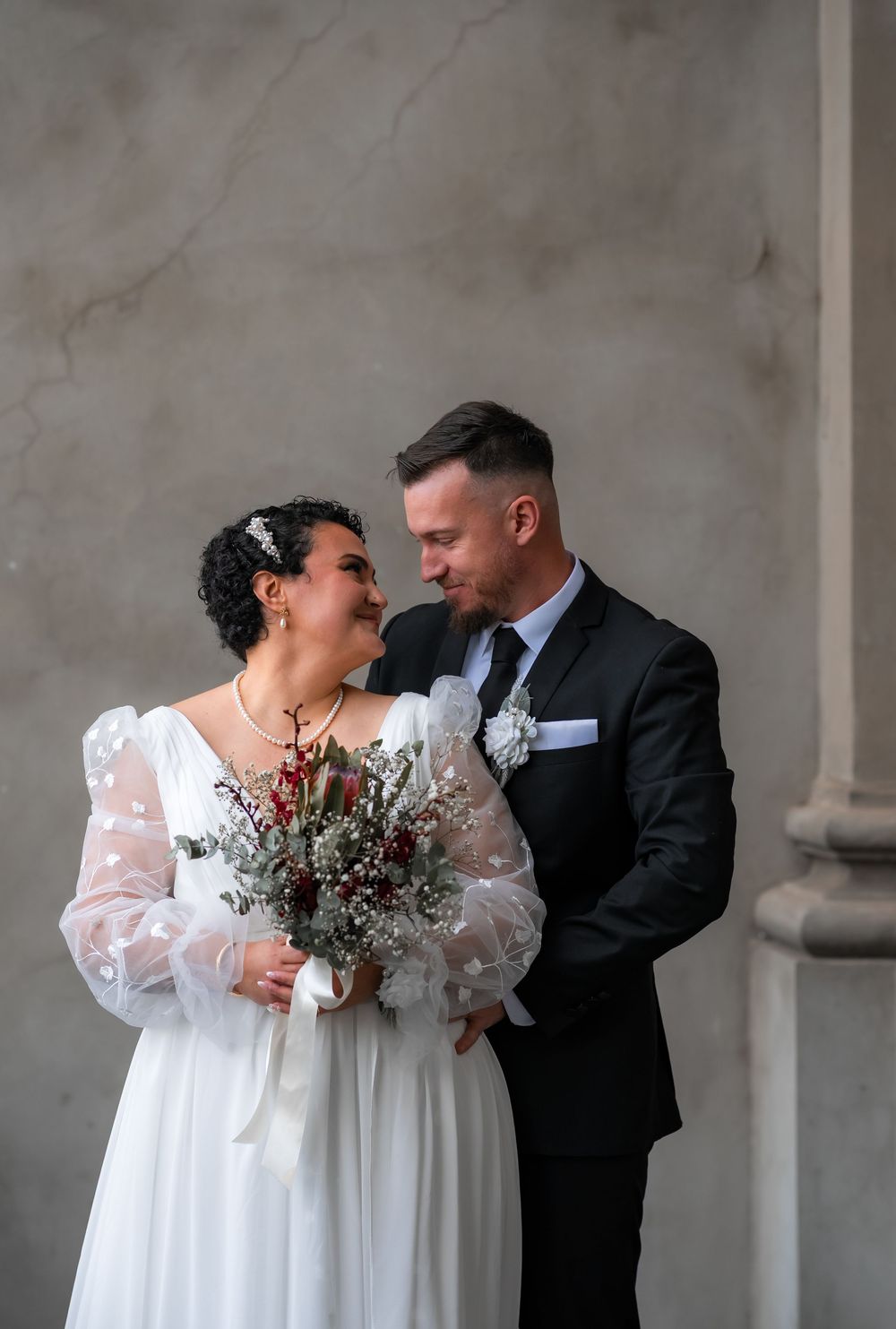 A couple in wedding attire share an intimate moment against a grey stone wall in a romantic outdoor setting.