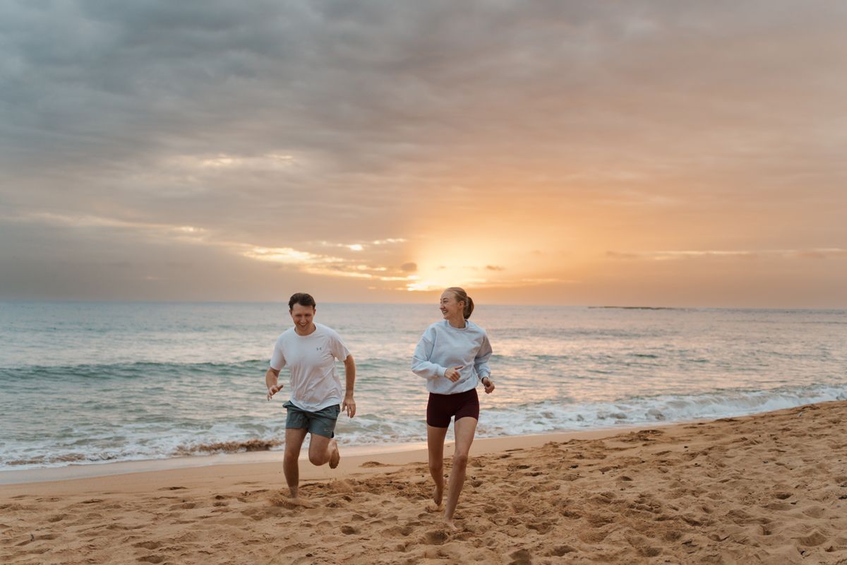 Two people running together on Newport beach at sunrise with ocean waves and cloudy sky in the background, engagement photoshoot in Sydney