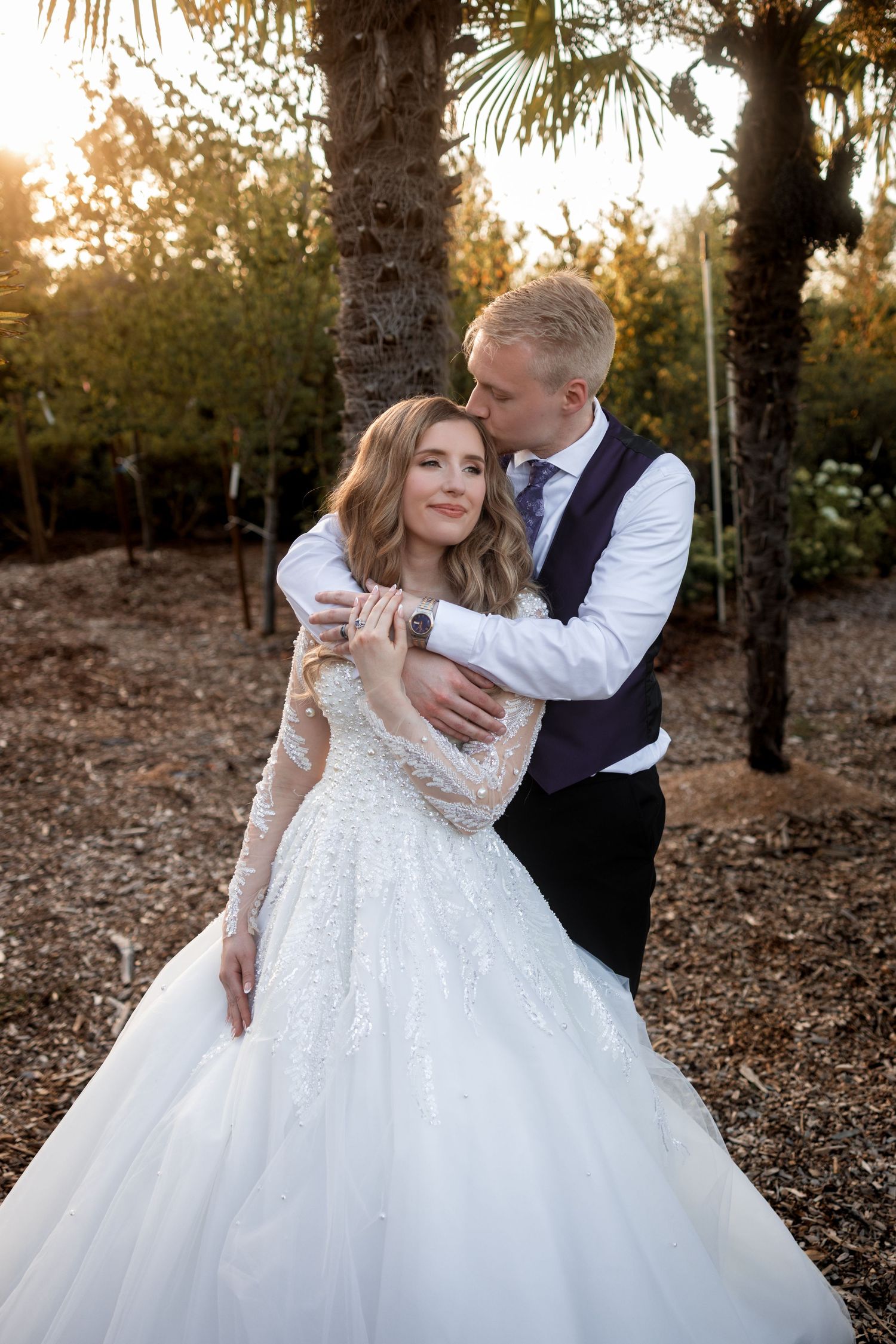 A romantic wedding photo of a couple embracing at sunset among palm trees with the bride wearing a lace white gown.
