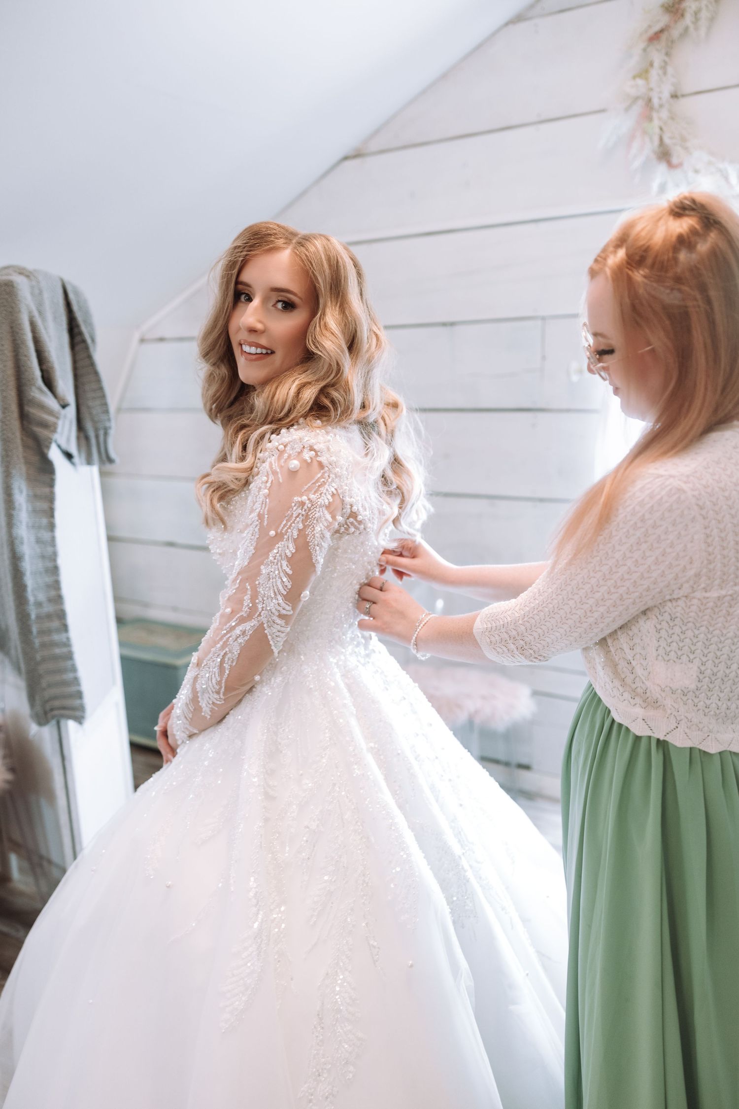 Bride in a sparkling white ballgown gets ready with help from attendants in sage green dresses.